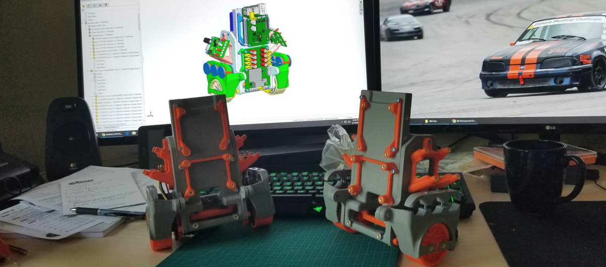 Two 3D-printed Bobble-Bot chassis on a cutting mat, with the CAD model on the monitor behind them